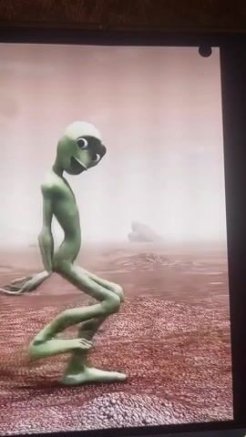 A vertical video taken close to a TV screen shows a digital animation of a green alien-like figure dancing. The figure is lanky with a large head, big eyes, and a wide smile. It moves energetically, bending its knees and changing its posture. The background is a muted reddish-brown landscape. Music plays audibly in the background.