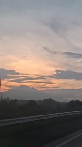 The video shows a landscape view from a moving vehicle, likely a car, as indicated by the motion blur and the presence of a guardrail in the foreground. The scene is dominated by a picturesque sunset or sunrise, with the sky displaying a gradient of colors, primarily soft oranges, yellows, and grays, interspersed with clouds. In the distance, a majestic mountain or a series of hills forms a silhouette against the vibrant sky. Below the mountains, a dark, hazy expanse suggests trees and vegetation, possibly fields, which are characteristic of a rural or semi-rural environment. The road surface, a dark gray asphalt, is visible in the foreground, suggesting the perspective of someone traveling on a highway or a similar thoroughfare. The overall atmosphere is serene and tranquil, capturing the beauty of nature during twilight. There are no people or distinct objects other than the natural elements and the road infrastructure visible.