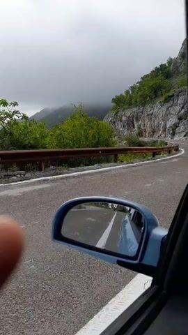 From inside a moving vehicle, the camera shows a view through the passenger window and side mirror, focusing on a winding mountain road. The road is flanked by a weathered metal guardrail and a steep, rocky hillside covered with sparse greenery.  The sky is overcast, with heavy grey clouds shrouding the mountain peaks in the distance.  The road surface is asphalt, and its curves are visible in the side mirror's reflection.  A thumb occasionally obstructs part of the foreground.  A cow is briefly seen on the side of the road.