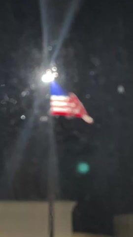 The video shows a blurred American flag waving on a pole, illuminated by a bright light source. The background is dark.