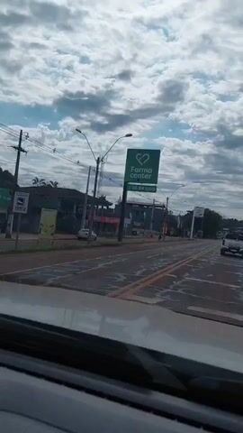 The video shows a view from inside a vehicle traveling down a street. On the left side of the street, there are buildings, including one with a green sign that says "Farma Center." There are also streetlights and power lines. The road has multiple lanes and appears to be asphalt with some cracking. There are other vehicles on the road, including a white truck and a white car. The sky is cloudy. On the right side of the street, there are more buildings and some people standing on the sidewalk. The video pans slightly to the right as the vehicle continues to move forward.