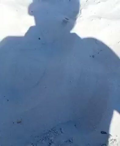 The video begins with a close-up shot of a person's shadow on a sandy beach. The shadow is in the foreground, and the sand is the dominant element in the frame. The perspective then widens to show a beach scene on a sunny day. In the mid-ground, a beach towel with a black cap and flip-flops is laid out on the sand. Further in the background, people are visible on the beach and in the water. Some are standing under umbrellas or tents, while others are swimming or wading in the lake. The water appears calm, and the sky is clear blue. The overall impression is a typical summer day at a crowded beach.