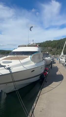 The video shows a waterfront scene with several boats docked at a pier. The camera pans from left to right, showcasing a large white yacht prominently in the foreground. It is moored to the pier, with ropes visible connecting it to the dock. Several black and red fenders hang from the side of the yacht to protect it from the dock. Further down the pier, other boats are docked, including a white speedboat and a sailboat with a tall mast. The boats are in calm, dark water, reflecting the sky and surrounding greenery. The background features a densely wooded hillside under a bright blue sky with scattered white clouds. The lighting suggests it is daytime, likely a sunny afternoon. No people are clearly visible interacting with the boats or the pier in the main focus of the video, although a person might be briefly glimpsed on one of the boats in the distance. The pier itself appears to be made of concrete and is casting shadows on the water. The overall atmosphere is peaceful and serene, depicting a typical marina setting.