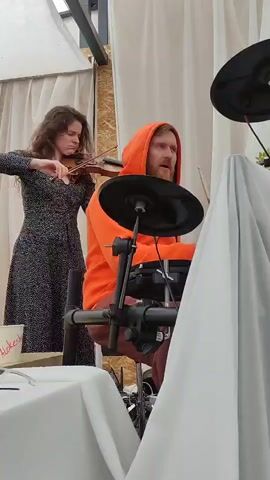 A woman with long, curly brown hair, wearing a black and white patterned dress, plays a violin. A man with a beard, wearing an orange hooded sweatshirt, sits behind an electronic drum set and plays it with drumsticks. They are both in what appears to be an outdoor setting, possibly under a canopy or tent, with draped white fabric visible in the background. A small table with white tablecloths is in the foreground.