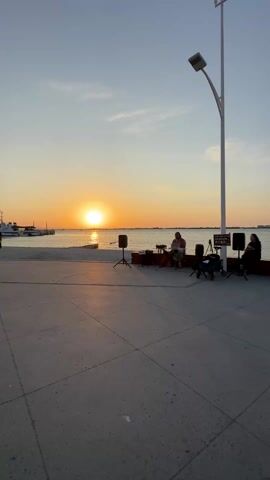 The video shows a band playing music on a pier at sunset. The sun is setting over the water, and there are boats in the distance. There are two speakers and a mixing board in front of the band. A woman sits next to the band, and a lamppost is behind them. The pier is made of concrete and is empty.