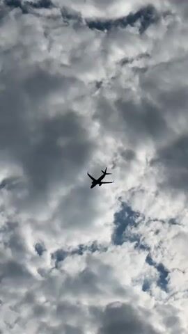 The video shows a silhouette of an airplane flying against a sky filled with clouds. The clouds are varied in density, with some appearing dark and others bright, suggesting a mix of light and shadow. The airplane is positioned in the center of the frame and appears to be moving. The camera angle is looking upwards towards the sky.