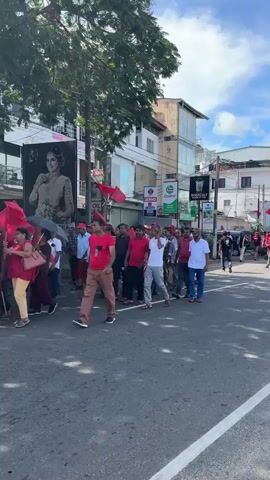 The video shows a crowd of people marching down a paved street. Many of them are wearing red shirts or hats and are carrying red flags. The street is lined with buildings, some of which have signs for local businesses, including a gas station and a restaurant. There is also a billboard featuring a woman in a dress. The sky is partly cloudy. The people appear to be peaceful and orderly.