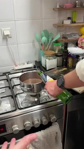 A person wearing a watch pours liquid from a green bottle into a silver pot on a gas stove. The stove is covered in aluminum foil and has four burners. A towel hangs from the oven door. The person's other hand rests on the counter next to the stove. Another person stands to the left of the stove. A counter is behind the stove with kitchen utensils and food ingredients on it. The wall behind the counter is tiled.
