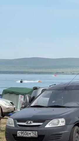 A group of people are enjoying a sunny day by a lake. In the foreground, a black car is parked, its license plate visible. Behind it, several tents and a light-colored car are set up, suggesting a campsite. The lake is active with a jet ski and an inflatable tube being pulled by a boat. In the distance, rolling hills covered in green vegetation form a scenic backdrop under a clear blue sky.