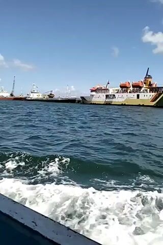 The video shows a view from a boat moving through the water, leaving a wake. In the background, several large ships are docked at a pier under a clear blue sky with scattered clouds. The ships appear to be passenger ferries or cargo vessels. Some have cranes visible. The water is a deep blue, and the waves are moderate.