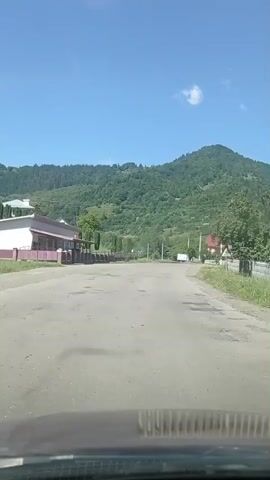 The video shows a view from the interior of a vehicle driving down a paved road. The road is bordered by a sidewalk and a building on the left and trees and houses on the right. In the background, there is a lush green mountain under a clear blue sky with a few scattered clouds. The vehicle is moving forward at a steady pace. The weather appears to be sunny and warm.