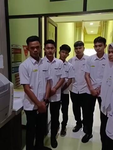 A group of seven young people, five males and two females, stand in a hallway. They are all wearing white shirts with badges on the left side of their chests and black pants. The females are also wearing white headscarves. The hallway has green walls and is well-lit. There are doorways visible on either side of the hallway. The people are looking towards the camera. The image is blurry, suggesting it was taken with a moving camera.