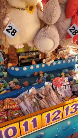 This is a close-up, low-angle, shaky shot of a carnival game booth. In the foreground, there are rows of prize dolls and bags of candy displayed on a shelf. Behind this, a blue trough is filled with colorful plastic fish. In the background, large stuffed animals, including a hedgehog, a teddy bear, and another fuzzy creature, are displayed, each with a numbered tag that reads "28." The environment appears to be outdoors, likely a fair or carnival, with bright lights and colorful decorations visible.