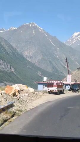 The video shows a view from inside a vehicle traveling along a mountain road. The road is paved and curves to the right. On the left side of the road, there is a steep, rocky slope leading up to a mountain. In the foreground on the left, there is a white truck with a red cargo bed and a blue and white building with a water tank on top. A black motorcycle is parked near the building. In the background, there are large, snow-capped mountains under a clear blue sky. The sun is shining, casting shadows and highlighting the rugged terrain. The vegetation appears sparse, with some trees scattered on the mountainside. The overall impression is of a remote, mountainous landscape.