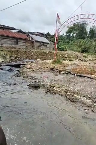 The video shows a first-person perspective of someone riding a motorcycle or similar vehicle through a shallow body of water that has flooded a dirt road. The water level is approximately halfway up the wheel of the vehicle. In the background, there are several simple wooden houses with corrugated metal roofs, situated on a hillside. The terrain appears to be rural and somewhat undeveloped, with loose rocks and debris along the roadside. The sky is overcast. There are brief glimpses of other vehicles and people in the distance, including a person on a motorcycle and a dark-colored car. The camera is positioned low, giving the impression of being close to the water's surface.