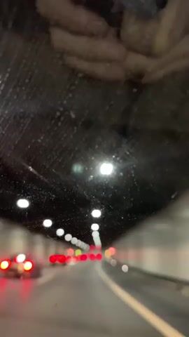 The video shows a point-of-view shot from inside a vehicle driving through a tunnel. The tunnel ceiling is dark and appears to have some textured material or discoloration. Lights are visible along the ceiling of the tunnel, spaced out at intervals. The road ahead is visible, with lane markings. Several vehicles are ahead, with their red taillights illuminated, indicating they are in motion or stopped. The windshield of the vehicle appears to have water droplets on it, causing some blurriness. The overall scene is dimly lit due to the tunnel environment.