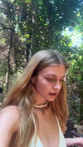A young woman with long, light brown hair walks through a tropical environment. She wears a white swimsuit top and a shell necklace. She looks slightly upwards, her mouth open in surprise or awe. The background consists of lush greenery with sunlight filtering through the leaves. The camera appears to be handheld, following her as she walks.