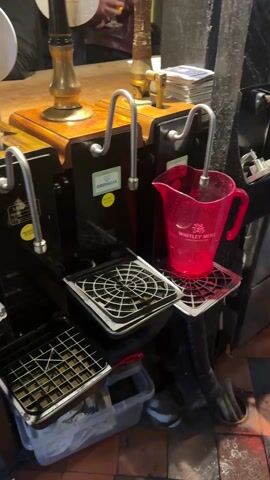 The video shows the behind-the-scenes area of a pub, focusing on the beer taps and the surrounding workspace. Several beer taps are visible, along with drip trays and containers beneath them. A red plastic pitcher sits on one of the drip trays. A variety of items, including beer cans, a bucket, a bottle, boxes, and glasses, are stored on shelves and the floor beneath the bar top. There is at least one person visible in the background near the taps. The floor is tiled with a red and brown pattern. Overall, the area appears cluttered and functional.