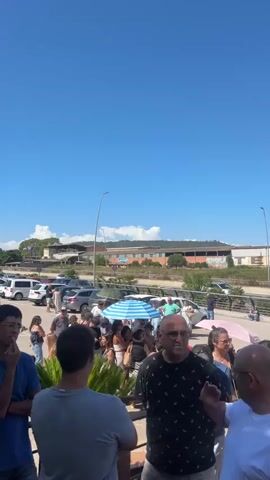 A group of people stand in a parking lot under a clear blue sky. The people are of various ages and genders, dressed in casual clothing. They appear to be talking and interacting with each other. In the background, there are several cars parked, and a building with a red roof. A blue and white striped umbrella is open amongst the crowd. The overall impression is of a casual outdoor gathering on a sunny day.