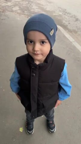 The media is a portrait of a young boy standing outdoors. He is wearing a blue knit beanie, a black puffer vest over a light blue long-sleeved shirt, and dark pants. He is also wearing dark sneakers with orange accents. The boy is looking directly at the camera with a slight smile. His expression is pleasant and engaging.

The setting appears to be an urban or suburban environment, possibly a street or sidewalk. The background is slightly blurred, but shows pavement, a curb, and some vehicles in the distance, suggesting a daytime scene. The lighting is natural and suggests it is daytime, likely overcast or in the shade given the soft lighting. There is a single yellow leaf on the pavement near the boy's feet.

The boy's attire suggests he is dressed for a cool or mild weather. The overall impression is a candid portrait of a child in a casual outdoor setting. There is no discernible text in the video.