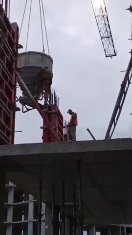 Two workers are on top of a concrete structure under construction. A crane is lowering a large bucket of cement. One of the workers is standing on a platform near the cement bucket, apparently preparing to release the cement into forms. The other worker, wearing an orange safety vest, is standing nearby. The sky is overcast.
