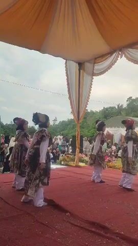 A group of four young girls in traditional attire are performing a dance on a red carpeted stage. The stage is covered by a large tent with a yellow and white canopy. The girls are wearing ornate dresses with floral patterns, white pants, and headscarves with matching embellishments. They are moving in unison, with their arms outstretched and hands gracefully posed. In the background, an audience of people are seated on the ground, watching the performance. The environment appears to be outdoors, with trees and a cloudy sky visible behind the tent.
