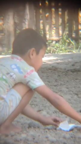 A young boy is crouched on the ground in an outdoor setting. He is wearing a light-colored t-shirt with a pattern of cartoon characters. He appears to be picking up small objects from the ground and putting them in his mouth. The ground is covered in small stones and dirt. Behind him, there is a wooden fence and some greenery. The lighting suggests it is daytime.