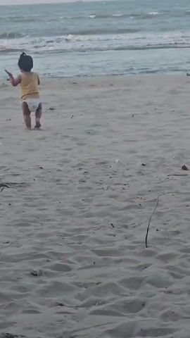 A young child wearing a diaper and a yellow tank top walks on a sandy beach towards the ocean. The child is facing away from the camera. The ocean has moderate waves and the sky is overcast. There is a plastic bottle on the sand to the left of the child.