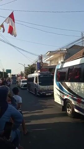 A street scene shows a procession of vehicles and people under a clear sky. A silver minibus with purple and gold accents is the most prominent vehicle, followed by other cars and buses. A red and white flag, possibly the Indonesian flag, hangs from a pole on the left. People are gathered on the side of the road, watching the procession. The environment appears to be a town or city with buildings and power lines lining the street. The sun is casting shadows, indicating it is daytime.