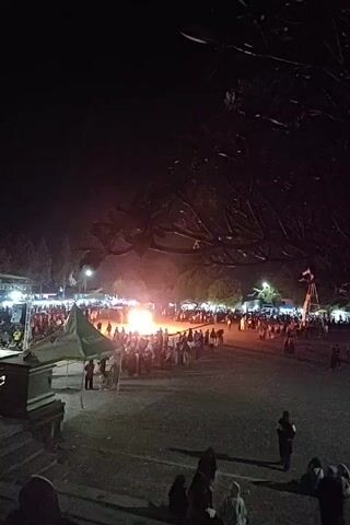 This video shows a nighttime outdoor gathering centered around a large bonfire. A crowd of people is standing around the fire, which is brightly lit and casts a strong orange glow. Tents and temporary structures, possibly stalls or stages, are visible on the periphery of the gathering, illuminated by lights. The ground appears to be an open, unpaved area. Trees with dark foliage are silhouetted against the night sky in the upper portion of the frame. The overall atmosphere suggests a festival or community event.