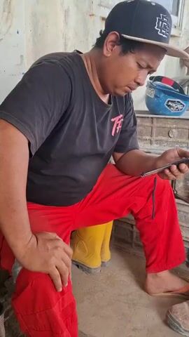 A man wearing a black t-shirt, red pants, a black baseball cap, and yellow boots is sitting down and looking at his phone. He is holding the phone in his right hand. His left hand is resting on his left knee. There is a blue hard hat on a shelf behind him. The environment appears to be outdoors, possibly a construction site, with a light-colored wall in the background. The lighting is bright.