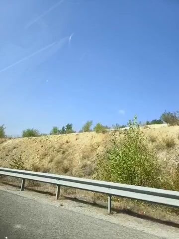 A video shows a view from a car window, looking out at a roadside embankment on a sunny day. The embankment is covered in dry, tan soil and rocks, with sparse green bushes and trees dotting the landscape. A metal guardrail separates the road from the embankment. The sky is a clear, bright blue with a few faint white wisps of clouds. The footage appears to be taken while the car is in motion.