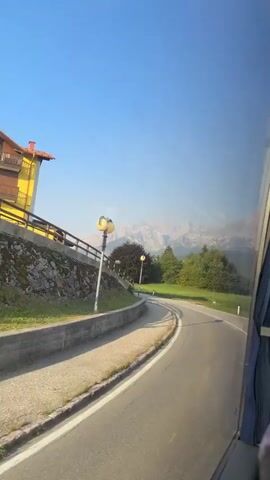 A vehicle, likely a bus or RV, is traveling on a road with a clear blue sky above. The road curves to the right, with a sidewalk and a stone retaining wall on the left. A yellow building with an orange roof is visible on the left side of the road, next to a wooden fence. Streetlights are present along the road. In the distance, mountains can be seen. Trees and other houses are scattered throughout the landscape. The right side of the video shows the interior of the vehicle, including a window with a blue curtain. There are no people clearly visible in the scene.
