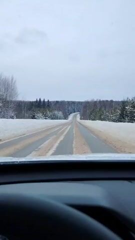The video shows a car's point of view driving down a snowy, paved road. There are snow-covered trees on either side of the road and a grey, overcast sky. The road is slightly uphill and winding. There are road signs on both sides of the road. The car's dashboard is visible at the bottom of the frame.