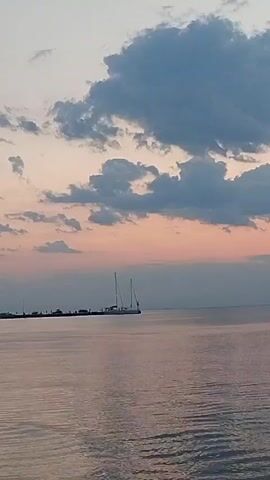 A slow-motion video clip captures a serene seascape at dusk. The camera pans slightly across a calm body of water, reflecting the soft hues of the sky. Dominating the mid-ground is a sleek white catamaran, its twin masts standing tall against the pastel-colored clouds. The catamaran is docked at a pier, where a few dark figures can be seen on the shore. The sky is a gradient of soft pinks and blues, transitioning into a deeper gray as it ascends. The overall atmosphere is one of tranquility and the fading light of day.