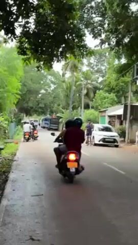 The video shows a street in what appears to be a tropical or subtropical area. The road is asphalt and has a drainage ditch on the left side. There are several trees and green plants on either side of the road. A number of motorbikes with one or two riders each are driving down the street. A white car is parked on the right side of the road. A man is standing near the car, and another man is walking on the left side of the road. A truck follows the bikes down the road.