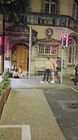 The video pans across a nighttime street scene in front of a building with "SUNLIGHT CHAMBERS" written above a large wooden door. Several people are sitting on the steps of the building. To the right, two men stand near a traffic light. Further down the street, a group of people stand outside a shop with a sign that reads "Di Fontanes New York Pizza." A rainbow-colored statue of a fox is visible on the left side of the frame. The scene appears to be lively with people congregating on the sidewalk and in front of the businesses. The lighting suggests it is evening or night.