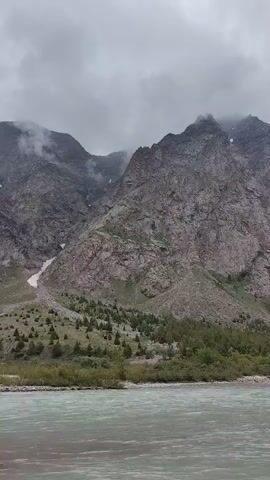 A wide, slow-moving river flows in the foreground under a cloudy sky. The water is a murky, pale green with visible currents and ripples. In the middle ground, a narrow strip of land separates the river from a steep, rocky mountain slope. Scattered evergreen trees dot the lower part of the mountain. The mountain itself is massive and rugged, with scree slopes and patches of snow or ice visible higher up. The peak is partially obscured by heavy clouds, giving the scene a somber, remote atmosphere.