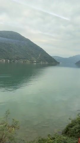 The video shows a view from a moving vehicle, likely a train, looking out at a large lake surrounded by green, rolling hills. The water is a vibrant turquoise color, and the sky is overcast with some streaks of light suggesting sunlight trying to break through. Lush green foliage lines the foreground of the video, indicating the vehicle is traveling along the shore of the lake. The hills on the opposite side of the lake are dotted with small buildings, possibly houses or villages. The overall impression is one of a scenic and peaceful landscape.