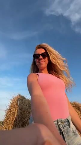 A young woman with long blonde hair and sunglasses is taking a selfie in a field of hay bales under a blue sky. She is wearing a pink tank top and jeans and smiling at the camera, with one arm extended. The background shows more hay bales and a distant landscape with trees and hills. The lighting suggests it is either early morning or late afternoon.