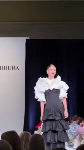 A model walks a runway at a fashion show. The model has their hair pulled back in a bun. They are wearing a black strapless top with a large white, ruffled collar. The skirt is tiered and ruffled as well, starting from the waist down to the floor. A crowd of people watches from the sidelines. The backdrop is a black curtain and a white wall with the word "RERA" visible. Spanish music plays loudly. The model turns to show the back of the outfit, and then continues walking off the runway.