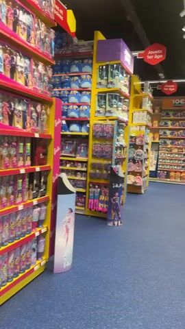 This media file captures a wide shot of a toy store aisle, likely in Clermont-Ferrand, France, given the context. The primary subjects are shelves brimming with a vast array of toys, predominantly dolls and play sets. The shelves are organized by color and type of toy, with bright pink and yellow units dominating the scene. The floor is a vibrant blue carpet, and the lighting suggests an indoor environment, likely during daytime business hours, given the commercial setting.

On the left side of the aisle, shelves are stacked high with dolls, many in individual boxes, and pink shopping bags adorned with unicorn and star motifs hang prominently. The dolls appear to be aimed at young girls, with many featuring princess-like attire. Further down, more shelves display various toy sets, possibly including doll accessories or play kitchens.

The right side of the aisle features more shelving units, also packed with toys. A prominent yellow ladder or scaffolding-like structure is positioned in the aisle, suggesting it's used for stocking high shelves. In the distance, more aisles of toys are visible, indicating a large store. A blue trash receptacle is also noticeable further down the aisle.

The overall impression is one of abundance and variety, catering to children's play and imagination. The bright colors and organized displays create a cheerful and inviting atmosphere typical of a toy store. There is no visible text that is clearly legible in the footage, apart from what appears to be brand logos on some packaging. The activity is that of a retail environment, with the implied event being the selling of toys. No people are clearly visible in the frame, focusing the attention solely on the merchandise.