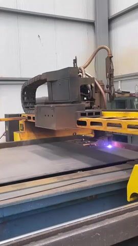 A plasma cutter is actively cutting through a sheet of metal. The cutting torch emits a bright blue and purple flame, accompanied by sparks. The plasma cutter machine itself is large and industrial, with a prominent track mechanism and a control unit. It is positioned over a flat metal surface, which appears to be on a larger machine bed with blue and yellow components. The environment is an indoor industrial space with white corrugated walls and a metal ceiling. The video captures the precise and intense process of metal fabrication.