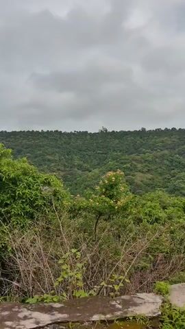 The video shows a landscape view of green hills under a cloudy sky. In the foreground, there is a concrete structure with a crack in it, and on the crack, there is a yellow and black object, possibly a traffic cone or a barrier. The camera pans from left to right, showing more of the lush green vegetation and rolling hills. The overall impression is of a natural, possibly mountainous, environment on an overcast day.