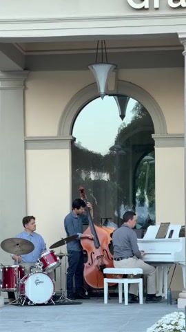 A jazz trio performs in front of a building with a large arched window. From left to right, the musicians are a drummer playing a red drum set, a bassist playing an upright bass, and a pianist playing a white grand piano. The drummer is wearing a blue plaid shirt and dark pants. The bassist is wearing a blue button-down shirt and dark jeans. The pianist is wearing a grey button-down shirt and grey pants. The scene appears to be outdoors during the day, with natural light illuminating the musicians and the building. The building has a light beige facade with white trim around the arched window. A modern-style light fixture hangs from the ceiling above the window. The reflection in the window shows trees and possibly other buildings.