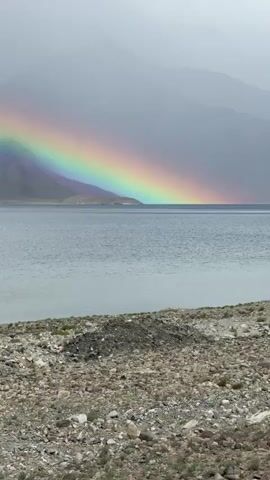 A static, eye-level, full-shot video displays a serene natural landscape. In the foreground, a rocky shoreline lines the bottom of the frame. Beyond it, a vast, calm body of water stretches across the middle ground. In the background, a mountain range is visible, partially obscured by fog and clouds, under a muted sky. A vibrant rainbow arches across the sky and appears to touch the distant mountains and the water. The video is completely silent.