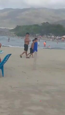 A man walks with two boys on a sandy beach. The man is wearing black shorts and no shirt. The boy in the middle is wearing a blue shirt and black shorts. The boy on the right is wearing black shorts. In the background, people are swimming and relaxing in the ocean and on the beach. Trees and mountains are visible in the distance. The sky is overcast.