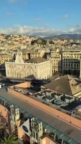 The video shows a panoramic view of a city with various buildings and a clear blue sky with some clouds. The city has a mix of old and modern architecture with buildings of different colors. There are palm trees lining a waterfront area and a road with cars moving along it. A building with the word "EATALY" is visible in the foreground. The video pans to reveal more of the city skyline and the surrounding hills.