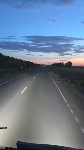 This video captures a nighttime drive on a paved road, likely in Baiqadam, Kazakhstan. The headlights of the vehicle are prominently illuminating the road ahead, creating a bright pathway through the twilight. The sky is a deep blue, transitioning to shades of pink and orange near the horizon, indicating either sunrise or sunset. Wispy clouds are scattered across the sky.

The road is bordered by grassy verges and trees on either side, with some distant hills or elevated terrain visible to the left. There are occasional signs or poles along the roadside, and a few vehicles, including what appear to be trucks, are visible in the distance, their taillights glowing red. The overall scene evokes a sense of a journey or transit during the transitional hours of the day. There is no visible text in the footage.