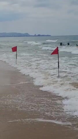 This media file captures a beach scene in Vityazevo, Russia, on an overcast day. The primary subjects are two red flags planted in the shallow water near the shoreline. These flags are likely warning signals, suggesting potentially hazardous conditions in the sea, such as strong currents or rough waves.

The scene is set at a beach with calm, sandy shores and moderately choppy water. Waves are breaking, creating foam and whitecaps that reach the flags. In the background, the sea extends towards the horizon, where a hazy silhouette of distant land and buildings can be observed. The sky is uniformly gray, indicating cloudy weather, and there are no distinct shadows, suggesting it might be morning or late afternoon.

There are no visible people in the frame. The only other discernible objects are the flags themselves, their poles, and a dark, indistinct mass at the base of the right flag, possibly a rock or some debris. The overall mood is somewhat somber due to the overcast sky, but the presence of the red flags implies an environment where safety is a concern, adding a sense of caution to the scene. No text is visible within the media file.